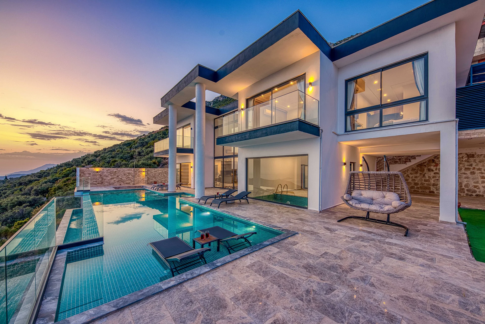 A modern luxury villa with illuminated glass-fronted rooms and an infinity pool overlooking the sunset